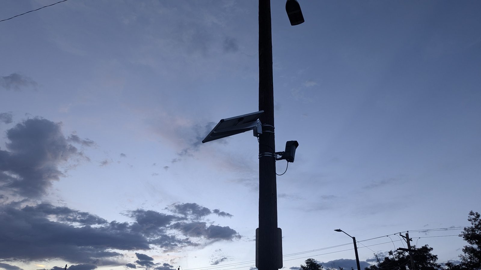 A pole-mounted roadside camera and solar panel overlook the street at dusk.
