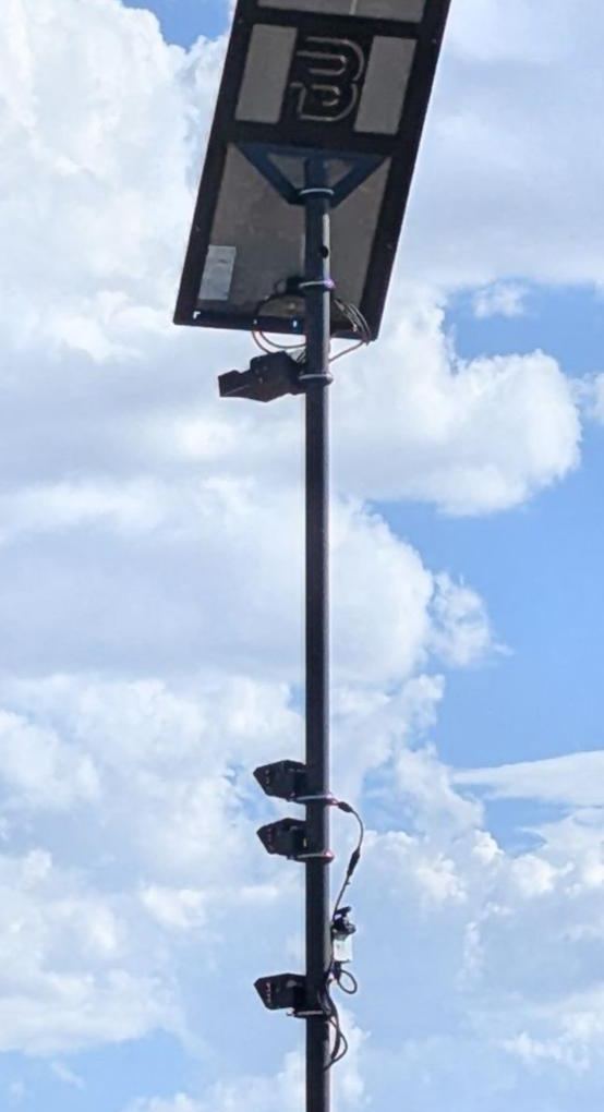 A tall solar camera tower with a stylized B on its panel and cameras stacked down the mast