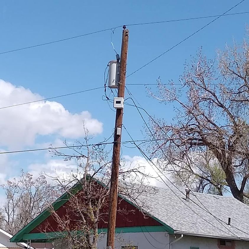 A white rectangular enclosure with a dark front panel mounted immediately below a cylindrical transformer on a wooden pole